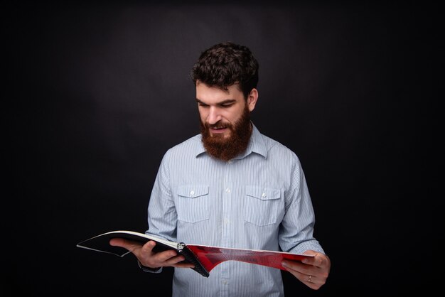 Junger bärtiger Mann, der in einem schwarzen Studio eine Zeitschrift oder ein Buch liest.
