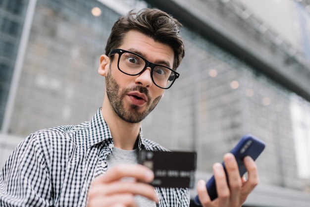 Junger bärtiger Mann, der Kreditkarte und Smartphone hält und Zahlung leistet. Hipster mit emotionalem Gesicht online einkaufen. Online-Banking-Konzept