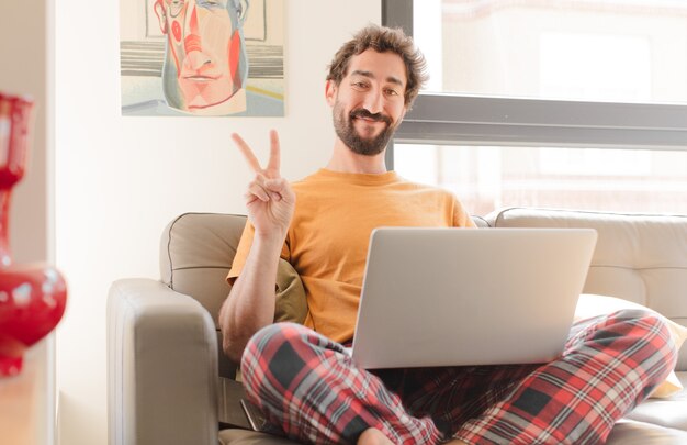 Junger bärtiger Mann, der lächelnd und glücklich sorglos und positiv gestikulierend Sieg oder Frieden mit einer Hand schaut und mit einem Laptop sitzt