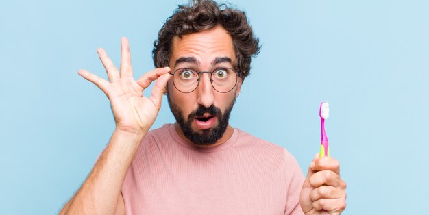 Junger bärtiger Mann, der sich schockiert, erstaunt und überrascht fühlt und eine Brille mit erstauntem, ungläubigem Blick hält