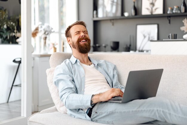 Junger bärtiger Mann, der zu Hause mit Laptop-Computer auf Sofa sitzt