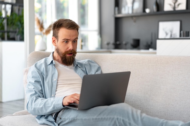 Junger bärtiger Mann, der zu Hause mit Laptop-Computer auf Sofa sitzt