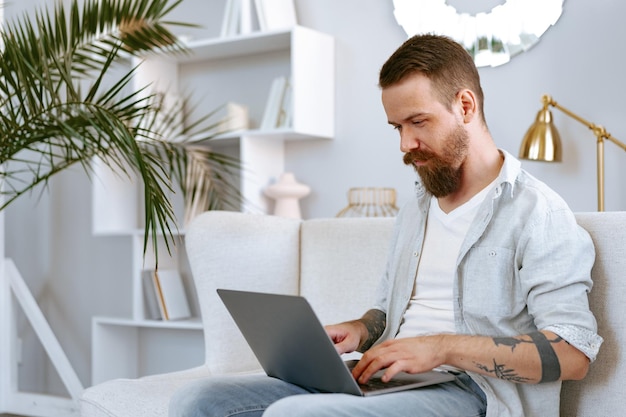 Junger bärtiger Mann, der zu Hause mit Laptop-Computer auf Sofa sitzt