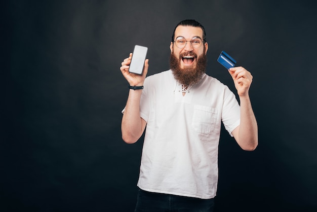 Junger bärtiger Mann freut sich über sein Internet oder mobiles Banking mit seiner neuen Karte und seinem Telefon.