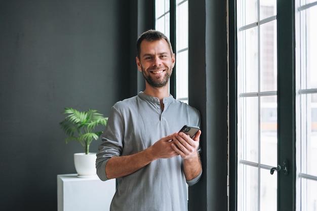 Junger bärtiger Mann in grauem Longsleeve mit Smartphone in den Händen, der eine Nachricht liest, die am Fenster im modernen Büro mit dunklen Wänden steht