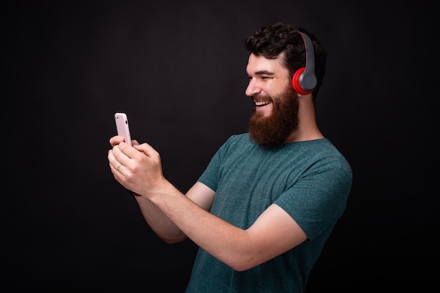 Junger bärtiger Mann macht ein Selfie und hört die Musik über rote Kopfhörer auf schwarzem Hintergrund.