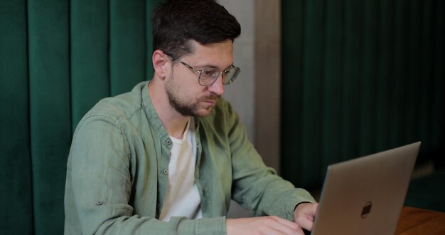 Junger bärtiger Mann mit Brille, der online remote im Café arbeitet Mann mit Computergeräten