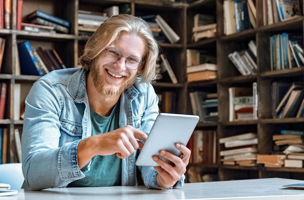 Junger bärtiger Mann mit Brille und Bibliothekstisch hält modernes Tablet in die Kamera und lacht