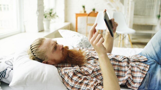 Junger bärtiger Mann mit Tablet-Computer, der zu Hause im Schlafzimmer auf dem Bett liegt