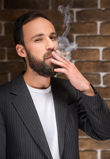 Junger bärtiger Mann raucht gegen Backsteinmauer.