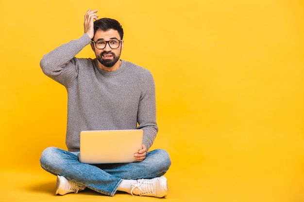 Junger bärtiger Mann schockiert überrascht erstaunt mit Laptop-Computer. Lustiges Bild des jungen kaukasischen männlichen Studentenmodells, das auf Boden sitzt