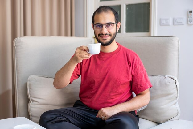 Junger bärtiger Mann trinkt Kaffee und lächelt im Wohnzimmer