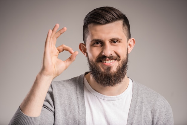 Junger bärtiger Mann zeigt okayzeichen auf grauem Hintergrund.