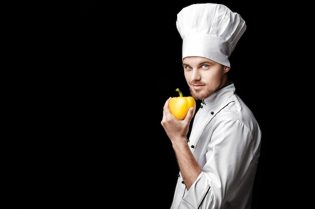 Junger bärtiger Mannkoch in weißer Uniform hält gelbe Paprika auf schwarzem Hintergrund