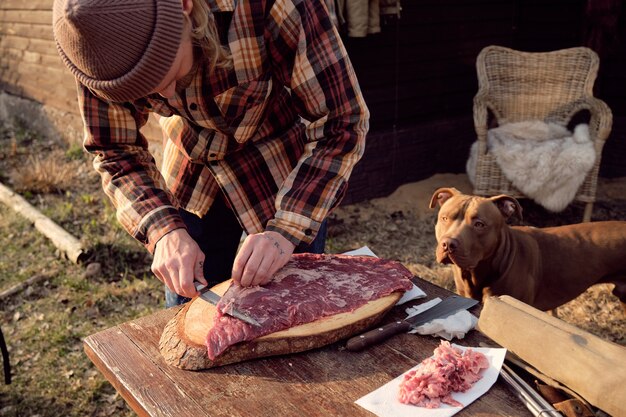 Junger Bauer, der frisches Fleisch zum Grillen schneidet, während sein Hund in der Nähe steht und auf den Prozess achtet