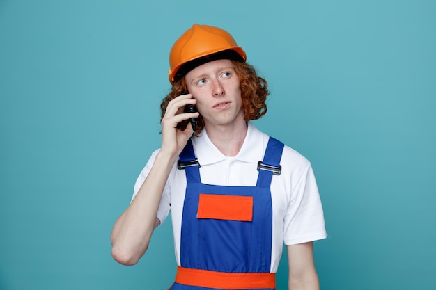 Junger Baumeister in Uniform spricht am Telefon isoliert auf blauem Hintergrund