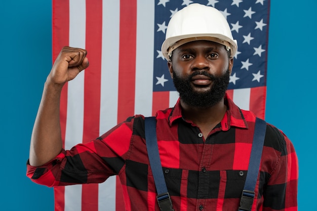 Junger Baumeister oder Vorarbeiter afrikanischer Abstammung in Arbeitskleidung und Helm zeigt seinen starken Arm im Ellbogen gegen die amerikanische Flagge