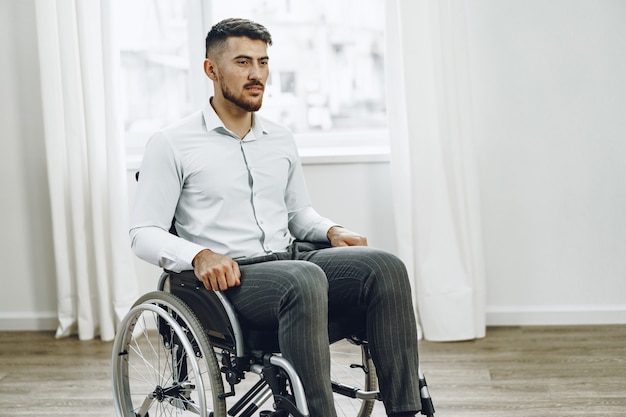 Junger behinderter Mann, der in einem Rollstuhl nahe dem Fenster zu Hause sitzt