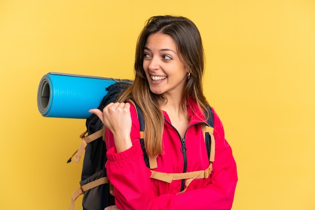 Junger Bergsteiger mit einem großen Rucksack isoliert auf gelbem Hintergrund, der auf die Seite zeigt, um ein Produkt zu präsentieren