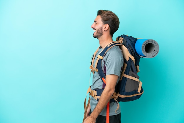 Junger Bergsteiger mit großem Rucksack isoliert auf blauem Hintergrund lachend in Seitenlage