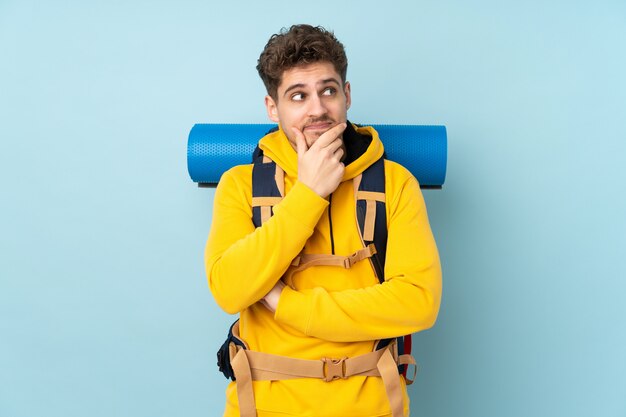 Junger Bergsteigermann mit einem großen Rucksack lokalisiert auf blauer Wand, die eine Idee denkt