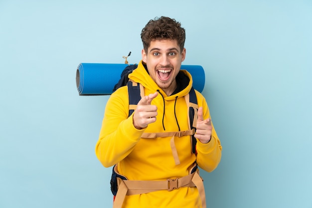 Junger Bergsteigermann mit einem großen Rucksack lokalisiert auf blauer Wand zeigt Finger auf Sie