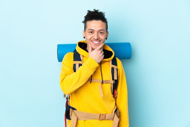 Junger Bergsteigermann mit einem großen Rucksack über isoliertem blauem Hintergrund lachend