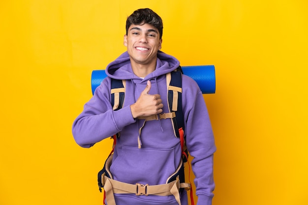 Junger Bergsteigermann mit einem großen Rucksack über isoliertem Gelb, der eine Daumen hoch Geste gibt