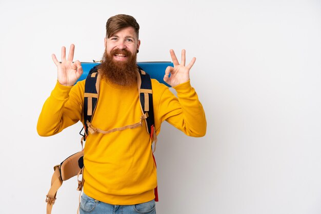 Junger Bergsteigermann mit einem großen Rucksack über isoliertem Weiß, das ein OK-Zeichen mit den Fingern zeigt