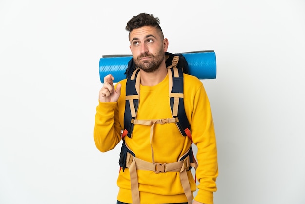 Junger Bergsteigermann mit einem großen Rucksack über isolierter Wand mit den Fingern, die kreuzen und das Beste wünschen