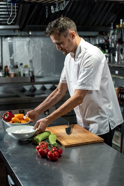 Junger Berufskoch des modernen Restaurants in weißer Uniform, der in der Küche am Tisch steht und Zutaten für die Gemüsegarnitur zubereitet