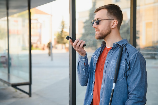 Junger blinder Mann mit Smartphone, der auf einer Bank im Park in der Stadt sitzt und ruft