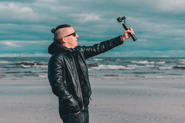 Junger Blogger in Sonnenbrillen, die Selfie machen oder Video am Strand mit Aktionskamera mit kardanischem Kamerastabilisator streamen.