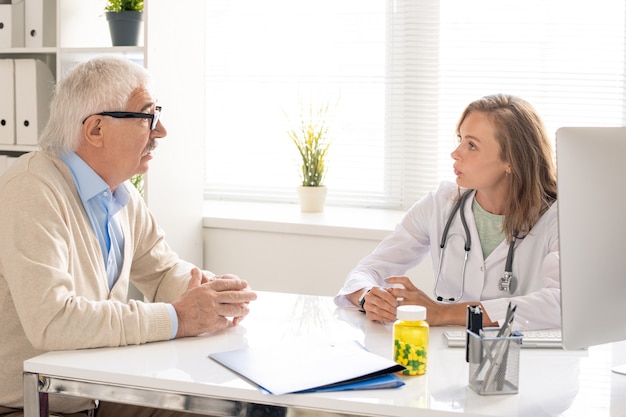 Junger blonder Arzt des medizinischen Beraters, der am Arbeitsplatz sitzt, mit dem älteren Patienten spricht und ihm einige Empfehlungen gibt