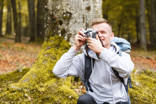 Junger blonder attraktiver lächelnder mann mit trekkingstöcken und kamera, der auf dem moos im wald sitzt und wunderbare waldansichten trekking-erholung und gesundes lebensstilkonzept aufnimmt