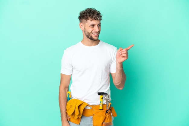 Junger blonder Elektriker, isoliert auf grünem Hintergrund, der mit dem Finger zur Seite zeigt und ein Produkt präsentiert