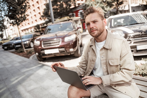 Junger blonder ernster Mann, der auf Bank mit Laptop sitzt, der online kommuniziert, Streaming, Videoanruf. Fernarbeit unterwegs. Lachender lächelnder hübscher Kerl. Stilvolles Millennial mit Social-Media-Netzwerk.