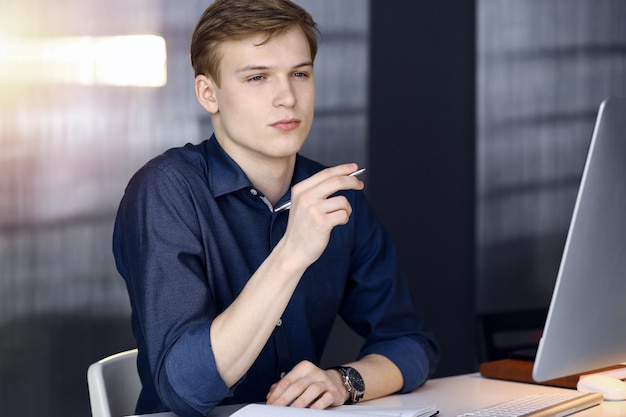 Junger blonder Geschäftsmann, der an seinem Arbeitsplatz mit Computer in einem abgedunkelten Büro über Strategie nachdenkt, grelles Licht im Hintergrund.