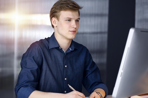 Junger blonder Geschäftsmann, der an seinem Arbeitsplatz mit Computer in einem abgedunkelten Büro über Strategie nachdenkt, grelles Licht im Hintergrund.