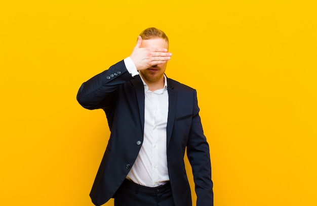 Junger blonder Geschäftsmann, der Augen mit einer Hand bedeckt, die sich ängstlich oder ängstlich fühlt, sich wundert oder blind auf eine Überraschung wartet