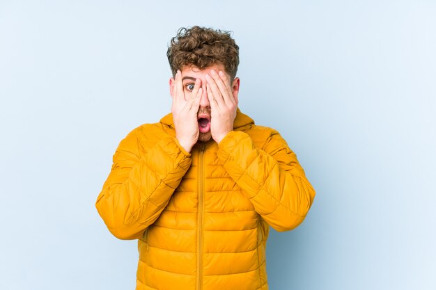 Junger blonder kaukasischer Mann des lockigen Haares blinzeln durch verängstigte und nervöse Finger.