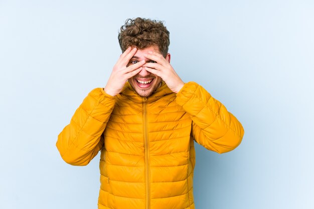 Junger blonder kaukasischer Mann des lockigen Haares isolierte Blinzeln durch Finger, verlegenes bedeckendes Gesicht.