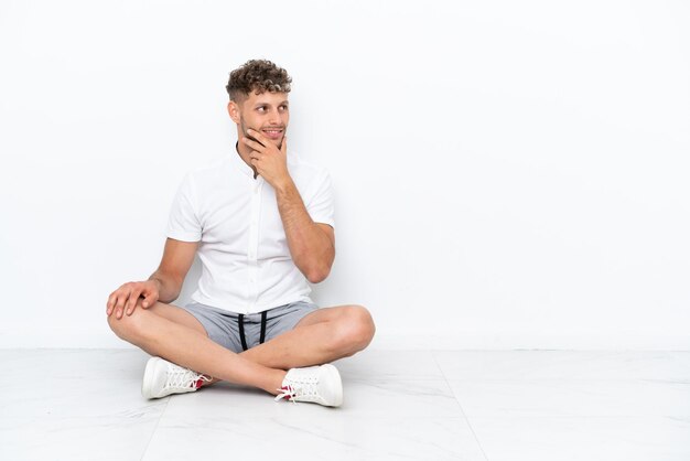 Junger blonder Mann, der auf dem Boden sitzt, isoliert auf weißem Hintergrund, hat Zweifel und einen verwirrten Gesichtsausdruck