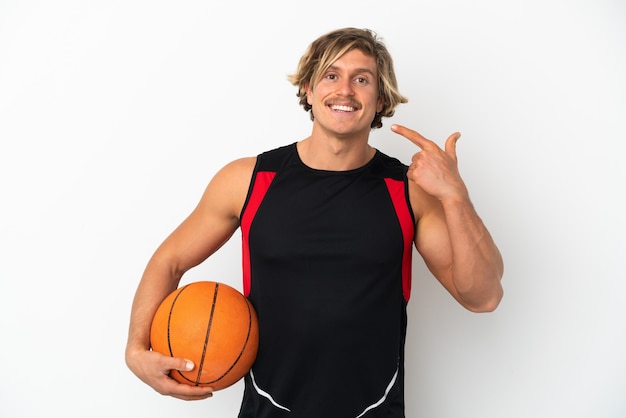 Junger blonder Mann, der einen Ball des Basketballs lokalisiert auf weißer Wand hält, die eine Daumen hoch Geste gibt