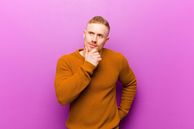 Junger blonder Mann, der einen Pullover trägt, der denkt, sich zweifelhaft und verwirrt fühlt, mit verschiedenen Optionen und sich fragt, welche Entscheidung zu treffen ist