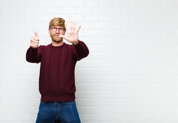 Junger blonder Mann, der freundlich lächelt und schaut, Nummer sechs oder sechst mit der Hand vorwärts zeigend, agaist Weinlesefliesenwand herunterzählend