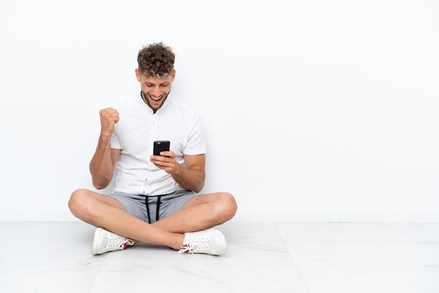 Junger blonder Mann, der isoliert auf weißem Hintergrund auf dem Boden sitzt, überrascht und eine Nachricht sendet