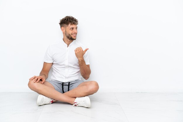 Junger blonder mann, der isoliert auf weißem hintergrund auf dem boden sitzt und zur seite zeigt, um ein produkt zu präsentieren