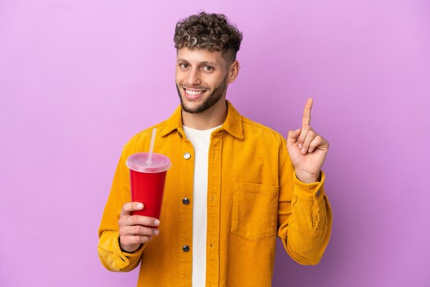 Junger blonder Mann, der Soda isoliert auf violettem Hintergrund hält und einen Finger im Zeichen des Besten zeigt und hebt