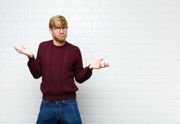 Junger blonder Mann, der verwirrt, verwirrt und gestresst aussieht, sich zwischen verschiedenen Optionen wundert, unsicheres agaist Vintage Fliesenwand fühlt